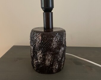 Puede incluir: Una lámpara de mesa de cerámica marrón con una superficie texturizada que se asemeja a la corteza de un árbol. La lámpara tiene un casquillo negro y un cable blanco.