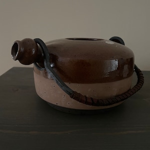 May include: A brown ceramic teapot with a black metal handle and spout. The teapot has a speckled glaze and a brown and black stripe around the middle.
