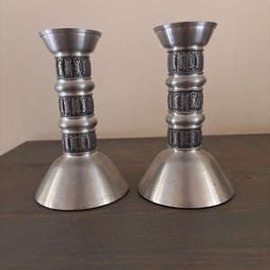 Puede incluir: Dos candelabros de metal plateado. Cada uno tiene una base ancha y acampanada, un tallo cilíndrico con detalles decorativos y una copa poco profunda en la parte superior. Los tallos presentan un patrón repetido de espirales y círculos. Los candelabros se exhiben sobre una superficie de madera oscura.