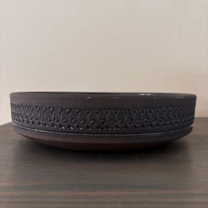 May include: A dark brown ceramic bowl with a decorative, repeating pattern around the rim. The bowl has a low profile and sits on a dark wooden surface. The rim design features a series of stylized shapes.