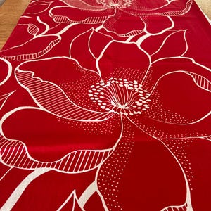 Op de afbeelding: Een rode tafelloper met een groot wit bloemenpatroon. De bloemen hebben gedetailleerde contouren en ontwerpen, wat een opvallend contrast creëert met de rode achtergrond. De loper is waarschijnlijk van stof.
