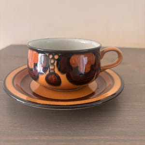 Peut inclure: Un ensemble tasse et soucoupe en céramique vintage. La tasse et la soucoupe présentent une palette de couleurs chaudes d'orange, de marron et de noir. La tasse a une forme arrondie avec une anse, et la soucoupe est circulaire avec des anneaux concentriques.