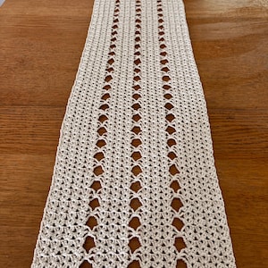 May include: A beige crocheted table runner with a delicate openwork pattern. The runner is laid out on a wooden table.