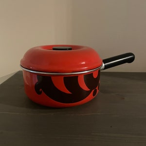 Puede incluir: Una cacerola de esmalte rojo con un diseño negro y un mango negro. La tapa tiene un pomo negro.