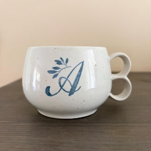 Op de afbeelding: Een witte keramische mok met een gespikkelde afwerking, voorzien van een blauwe cursieve letter A en een klein blauw takje met blaadjes. De mok heeft een opvallend handvat dat bestaat uit twee gestapelde cirkelvormige lussen.