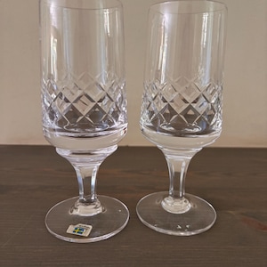 May include: Two clear crystal champagne flutes with diamond-cut patterns on the bowl. The flutes have a slender stem and a round base. A small label with a blue and yellow design is visible on the base of one flute.