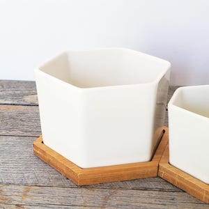 May include: Two white ceramic planters with a geometric design sit on square bamboo trays. The planters have a modern, minimalist aesthetic, perfect for small plants or succulents. The natural wood of the trays contrasts with the white of the planters.