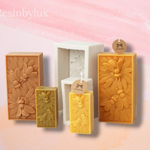 May include: Four silicone moulds for making candles, with three finished candles in the moulds. The candles are yellow and feature a design of bees and flowers. The moulds are white and have a rectangular shape. The candles have "Handmade" tags attached.