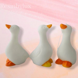 May include: Three white ceramic goose figurines with orange feet and beaks. The geese are facing different directions.