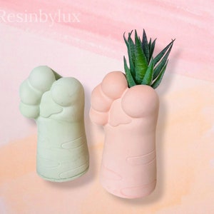 May include: Two paw-shaped planters, one green and one pink, with a small succulent plant in the pink planter.  Resinbylux is written in the top left corner of the image.