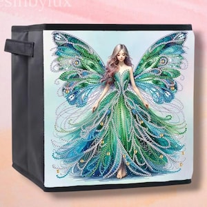 Peut inclure: Cube de rangement noir avec une illustration colorée d'une fée aux ailes et à la robe vertes et bleues, inspirées du paon. La fée a de longs cheveux ondulés et est ornée de bijoux. Le texte "Resinbylux" est visible.