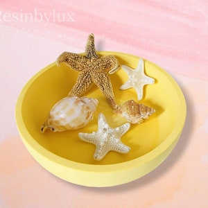 May include: A yellow bowl with a white rim filled with seashells and starfish. The bowl is on a pink and orange background.