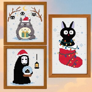 May include: Three framed cross-stitch artworks featuring characters in holiday settings. One shows a gray character with a Santa hat holding a gift. Another depicts a black cat in a Christmas stocking. The third shows a character holding a snow globe.