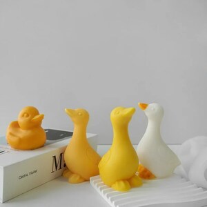 May include: Four decorative duck figurines. Three are yellow and one is white. The ducks are arranged on a white surface with a book in the background.