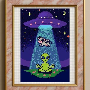 Op de afbeelding: Ingelijste kruissteekkunst met een gepixeld tafereel van een UFO die een koe omhoog straalt. Een alien mediteert eronder. De achtergrond is een sterrenhemel met een maan en roze bloemen.