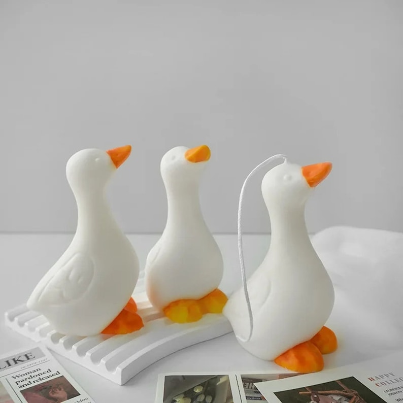 May include: Three white goose-shaped candles with orange beaks and feet. The candles are arranged on a white surface.