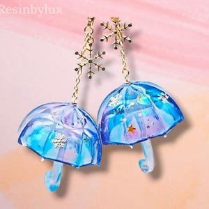 Può includere: Un paio di orecchini pendenti con ombrelli in miniatura in resina blu e viola con ciondoli a forma di fiocco di neve dorati. Gli ombrelli contengono stelle e glitter incorporati, sospesi da catenelle dorate e perni a forma di fiocco di neve.