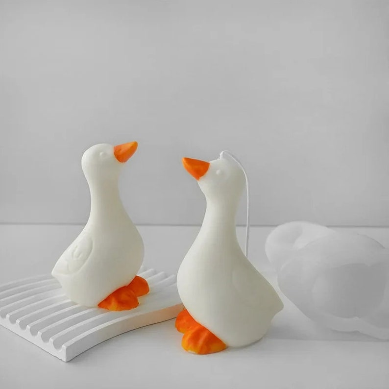 May include: Two white goose-shaped candles with orange beaks and feet. The candles are on a white surface.