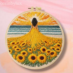May include: A circular embroidery hoop containing a textile art piece depicting a person with long dark hair standing in a field of yellow sunflowers. The person wears a long yellow dress that blends into the flowers. The background shows a blue ocean and a yellow sun rising in the sky.
