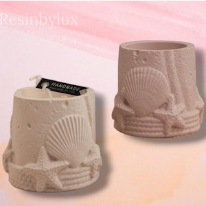 May include: Two off-white, cylindrical candle holders with seashell and starfish designs. One has a lit candle. The text "HANDMADE" is on a tag. The background is a soft pink and white.