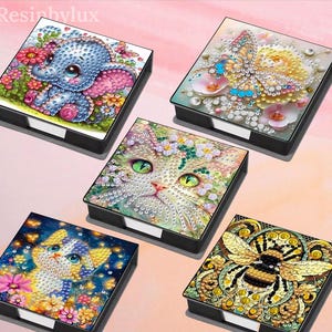 Peut inclure: Collection de cinq boîtes décoratives carrées à cadre noir. Chaque boîte présente un motif coloré et détaillé : un éléphant, un papillon, un chat, un chaton et une abeille. Les motifs sont réalisés avec de petits éléments scintillants, créant un effet mosaïque.