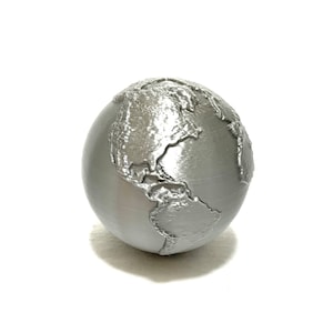 Puede incluir: Un globo de color plateado con un mapa del mundo en relieve texturizado. El globo es un modelo 3D impreso de la Tierra.