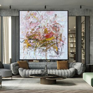 May include: Abstract painting with pink, white, and gold paint splatters and brushstrokes. The painting is framed and hanging on a wall in a modern living room.
