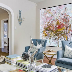 May include: A large abstract painting in shades of pink, white, and gold hangs on a white wall in a living room. Two blue armchairs with white pillows are positioned in front of the painting. A coffee table with books and a decorative sculpture sits in front of the chairs.