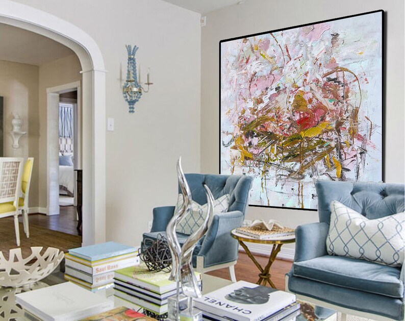 May include: A large abstract painting in shades of pink, white, and gold hangs on a white wall in a living room. Two blue armchairs with white pillows are positioned in front of the painting. A coffee table with books and a decorative sculpture sits in front of the chairs.
