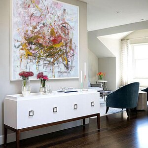 May include: Abstract painting in shades of pink, white, and gold with a white frame. The painting is hanging above a white dresser with four drawers and brown legs. The dresser is in a room with hardwood floors and a blue armchair.