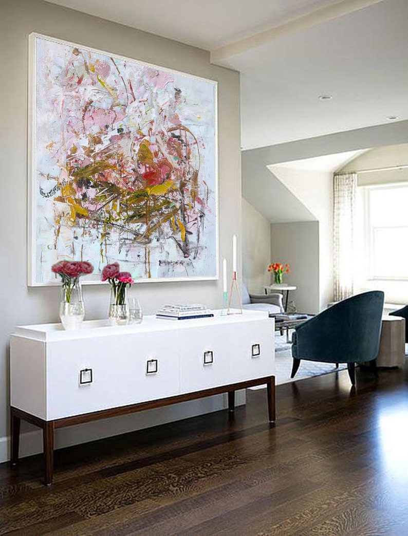 May include: Abstract painting in shades of pink, white, and gold with a white frame. The painting is hanging above a white dresser with four drawers and brown legs. The dresser is in a room with hardwood floors and a blue armchair.