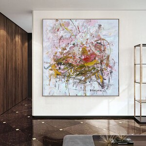 May include: Abstract painting with pink, white, and gold paint splatters and brushstrokes. The painting is framed and hanging on a white wall in a modern interior.
