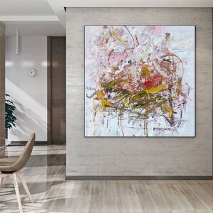 May include: Abstract painting with pink, white, and gold paint splatters and brushstrokes. The painting is framed and hanging on a wall in a modern home.