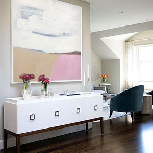 May include: A white, beige, and pink abstract painting hangs above a white dresser with four drawers. The dresser has brown legs and is in a room with hardwood floors.