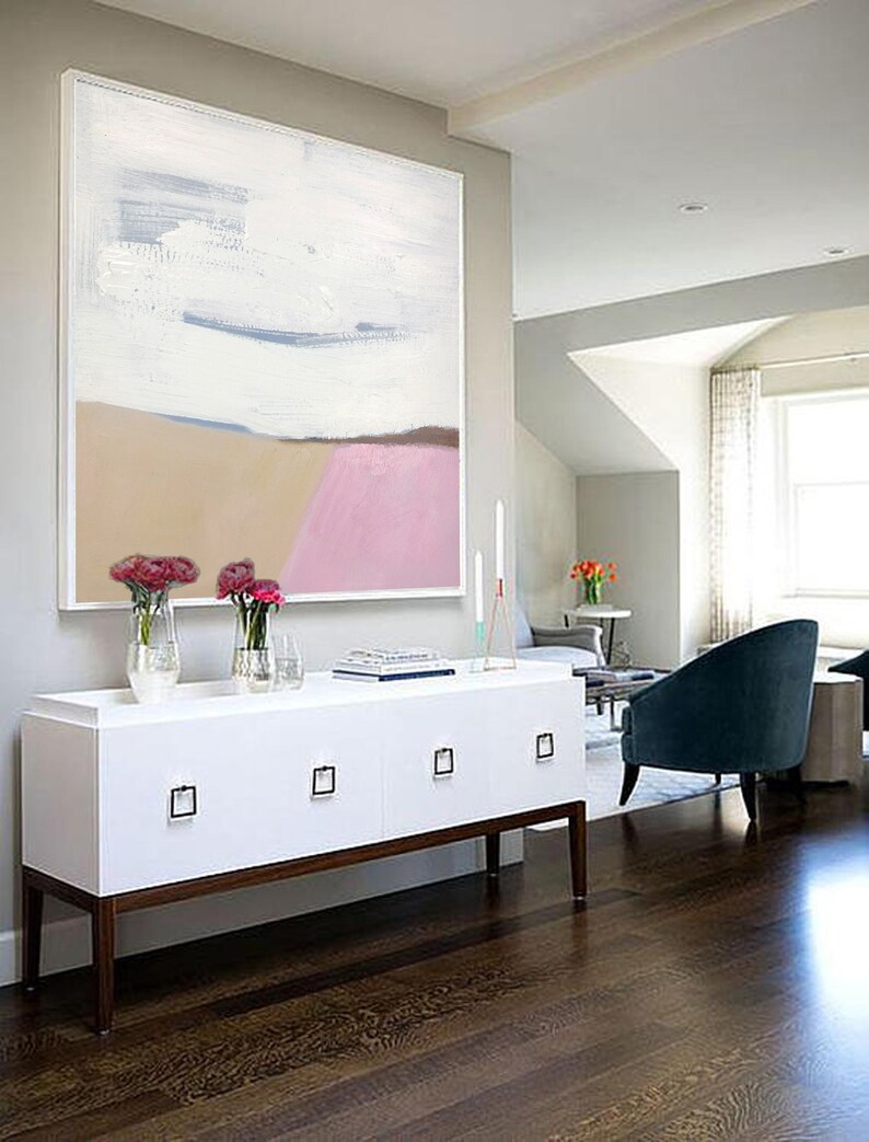 May include: A white, beige, and pink abstract painting hangs above a white dresser with four drawers. The dresser has brown legs and is in a room with hardwood floors.