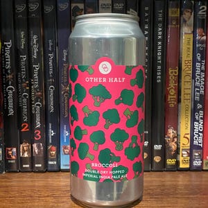 May include: A silver beer can with a pink label featuring green broccoli illustrations. The label reads "OTHER HALF" and "BROCCOLI DOUBLE DRY HOPPED IMPERIAL INDIA PALE ALE". The can is displayed in front of a shelf of DVDs.