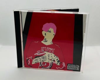 May include: A CD case featuring a stylized illustration of a person with pink hair and tattoos, wearing a red jersey with "NEW JERSEY DEVILS" text. The background is red with a Parental Advisory warning.