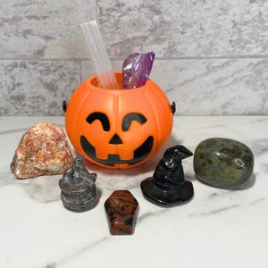 May include: An orange plastic jack-o'-lantern bucket with a smiling face, holding a clear crystal and a purple crystal. Surrounding the bucket are various stones and a small black witch hat, perfect for Halloween.