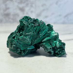 May include: A vibrant green malachite mineral specimen. The crystalline structure displays a radiating, fibrous pattern. The stone has a rough, textured surface and varying shades of green, from deep emerald to lighter tones.