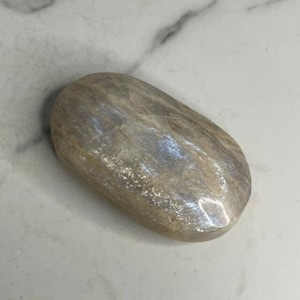 May include: A smooth, oval-shaped, light gray gemstone with iridescent shimmer. The stone has a natural, polished finish.