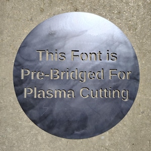 Pode incluir: Placa de metal circular com o texto "This Font is Pre-Bridged For Plasma Cutting" recortado. A placa tem uma aparência escura e manchada com um efeito de gradiente, e está sobre uma superfície de concreto.