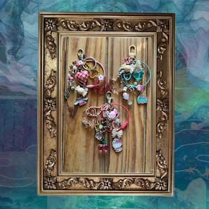 Pode incluir: Três chaveiros decorativos com vários berloques, incluindo corações, laços e figuras de personagens, são exibidos em um fundo de madeira dentro de uma moldura dourada ornamentada. Os chaveiros apresentam contas rosa, brancas e transparentes.