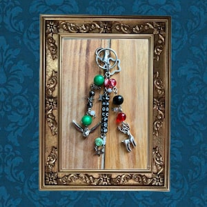 May include: A decorative keychain with a silver-tone ring and various charms. The charms include green and black beads, a deer, an anchor, and a charm that spells out "JEGULUS". The keychain is displayed against a wooden background within a gold-colored frame.