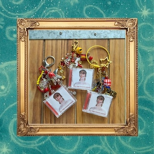 Puede incluir: Una colección de llaveros con imágenes de David Bowie en estuches de plástico transparente. Los llaveros están adornados con varios abalorios, cuentas y un acento en forma de estrella. El texto "ALL THE YOUNG DUDES" es visible en las carcasas de los CD.