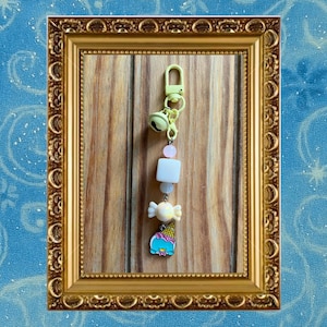 May include: A yellow keychain with a bell, beads, and a charm shaped like an ice cream cone. The keychain has a lobster clasp and a series of beads in white, pink, and light blue. The ice cream cone charm is blue, pink, and yellow.