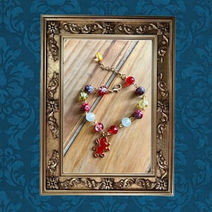 May include: A colorful beaded bracelet with red, purple, green, and white beads. The bracelet has a gold chain and a red charm. The bracelet is displayed inside a gold ornate picture frame on a wooden surface.