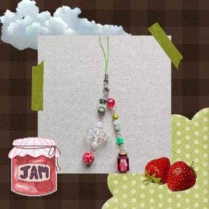 May include: A beaded charm with a green cord, featuring colourful beads in red, green, and clear. A small glass bottle charm with a strawberry design hangs below. The image also includes a strawberry jam jar illustration and two fresh strawberries.