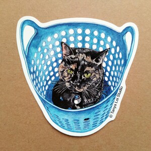 May include: A sticker featuring a tortoiseshell cat inside a blue laundry basket. The cat has green eyes and a collar with a tag. The basket has a perforated design and handles. The sticker is on a brown background.