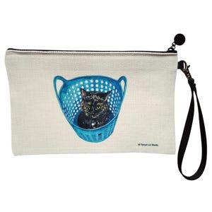 Puede incluir: Bolsa con cremallera de color crema con una ilustración en acuarela de un gato negro y marrón dentro de una cesta de lavandería azul. La bolsa tiene una correa de muñeca y un tirador de cremallera negros.
