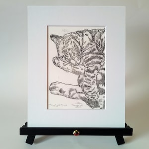 May include: A black and white monotype print of a tabby cat curled up while grooming, presented in a white mat board. The print features the handwritten text Monotype Print and a signature by Tanya Lai dated 2026 at the bottom of the artwork.
